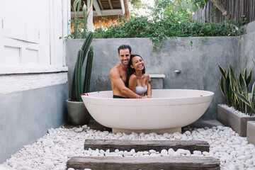 Happy young married enjoying recreation on resort during honeymoon taking bath together looking at camera, smiling woman and man in love having romantic spa therapy sitting in tub with rose petals