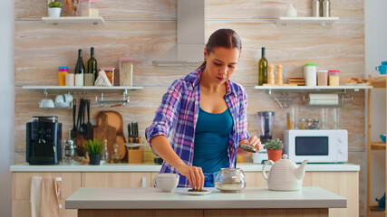Fototapeta premium Young woman preparing green tea for breakfast in a modern kitchen using teapot sitting near the table. Putting with hands, pressing herbal, healthy, tea leaves, in pot, in the morning.