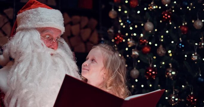 a little girl sits on the lap of Santa Claus and he reads her stories from a book
