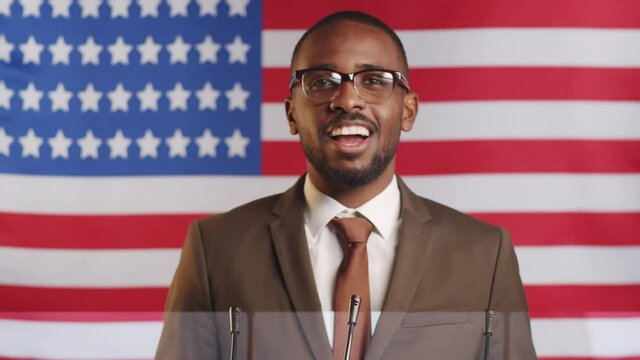 Young afro-american male politician in formal suit and glasses standing against U.S. flag and speaking into microphones while giving public announcement during press conference