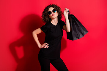 Photo of cute adorable curly woman wear casual black outfit eyeglasses holding bags sending kiss isolated red color background
