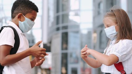 Multiethnic schoolchildren in safety mask disinfecting hands with sanitizer. The kid are standing at skyscrapers background .