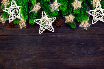 Fir branches, wicker stars and garland with warm lights on a dark brown wooden board. Christmas border festive background, traditional decorative ornaments. Copy space.