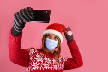 Fototapeta na wymiar A young girl in santa claus hat and medical protective mask takes a selfie on her phone camera. Congratulations during self-isolation, coronavirus pandemic. Selective focus