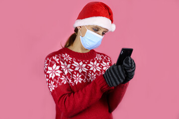 Fototapeta na wymiar A young girl in santa claus hat and medical protective mask looks into a mobile phone