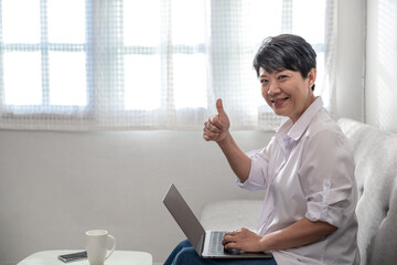 An enthusiastic senior woman raising her thumb up And working online from the laptop sitting on the couch at home. Home office ideas.