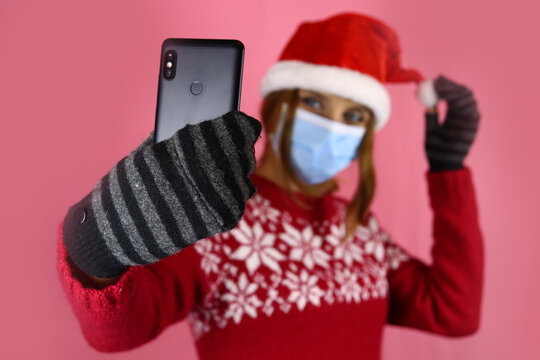 A young girl in santa claus hat and medical protective mask takes a selfie on her phone camera. Congratulations during self-isolation, coronavirus pandemic. Selective focus