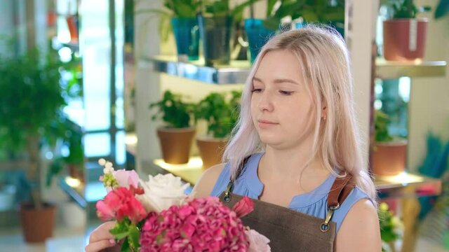 Young flower shop assistant with loose fair hair holds modern bouquet and looks at camera smiling gently closeup slow motion