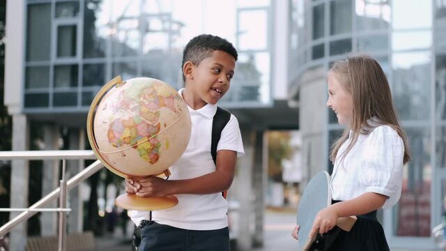 New school year. Elementary school. Two schoolkids holding hands are goig to school with a globe and bakpacks. City background