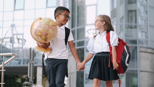 New school year. Elementary school. Two schoolkids holding hands are goig to school with a globe and bakpacks. City background