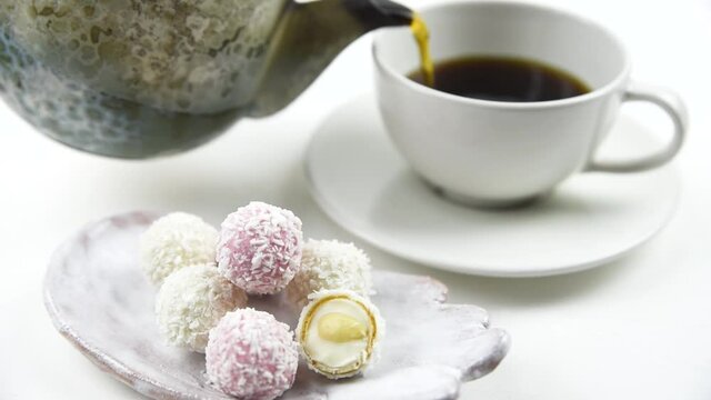 White coconut candy balls on handmade plate. Coconut cookies on white background with the cup of tea and teapot. Black tea pouring from the kettle. 