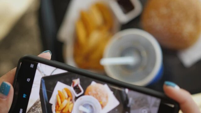 Woman using phone to take pictures of Burger, french fries and cola. Tourist girl have brunch or lunch time at fast food cafe. Top view