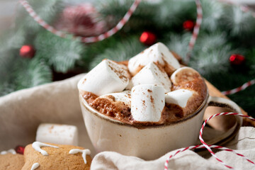 Hot cocoa or hot chocolate with marshmallows in a ceramic cup surrounded by gingerbread, Christmas tree branches and decorations on a brown wooden table