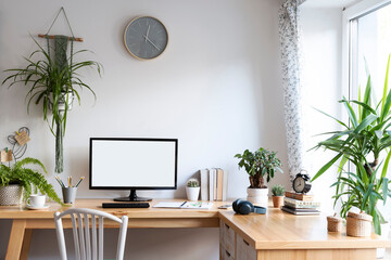 Modern interior of workplace at home with office desk and chair with blank white wall and window. Blank screen of computer with mock up, plant, clock and coffee. Mock up.