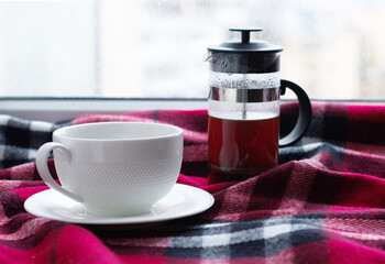 a pair of coffee, a saucer and a cup of coffee, next to a French press, stand on the windowsill, on a red, warm scarf. A cozy morning, autumn, a charge of cheerfulness, a single tea party. Divorce,