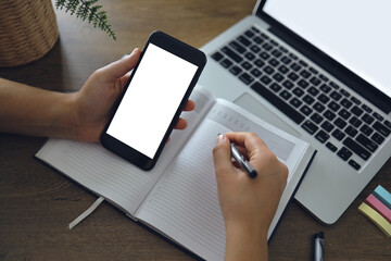 Mockup image of woman hand holding blank white desktop screen smartphone with laptop on table while writing her idea on notebook.