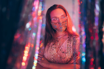 Christmas party look. Covid-19 fashion. Corporate celebration. Glamorous woman in eyeglasses festive dress designer golden chain crystal beads face mask neon light sparkle blur interior curtain.
