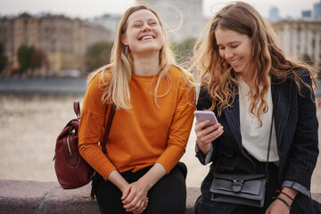 Two beautiful young woman friends walking together around the city, using a smartphone and having...