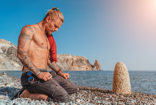 Mature Tattooed Man sitting on the beach before rock and pray. Ready To Fight on the sea background - Portrait Of A Handsome Muscular Ancient Warrior. Martial art concept