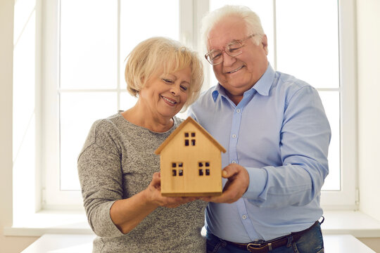 Smiling senior couple holding miniature house standing by the window in their new home