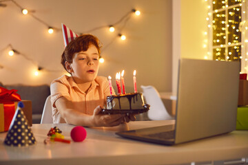 Little birthday boy making a wish and blowing burning candles on cake during online party at home