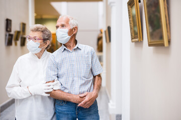 mature couple in mask protecting against covid examines paintings on display in hall of art museum