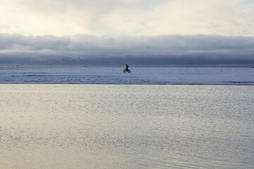 Fototapeta premium A man on a motorcycle rides through the snow on the river bank. Early winter. A motorcyclist rides on the snow cover of the shore of a reservoir, waves in cold water.