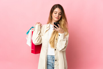 Young woman with shopping bag isolated on pink background holding shopping bags and writing a message with her cell phone to a friend