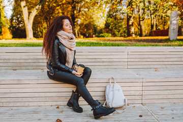 Positive afro hair woman wearing knitted sweater and scarf sit on bench in autumn park on sunny day with coffee and breathing deep