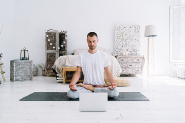 Calm caucasian male in casual wear with closed eyes meditating while listening online lesson using technology at home, tranquil spiritual man in lotus pose starting yoga video practice online
