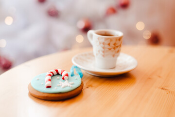 Cup of coffee with ginger bread cookie on the table near the white Christmas tree with golden bokeh lights