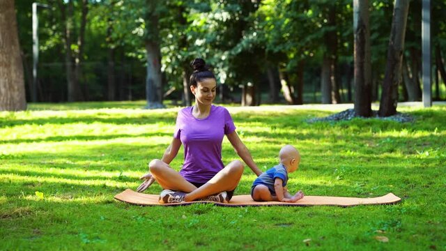 Fit woman in sportswear exercising on yoga mat in park, baby sitting beside. Fit mother and little child at outdoor training together. Concept of sport