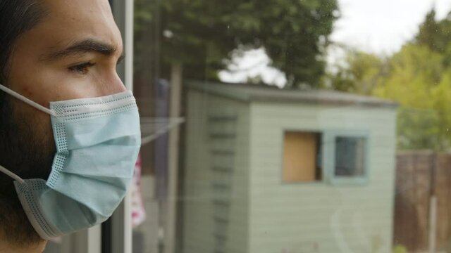 Male looking out of a window longingly wearing a facemask in quarantine close up
