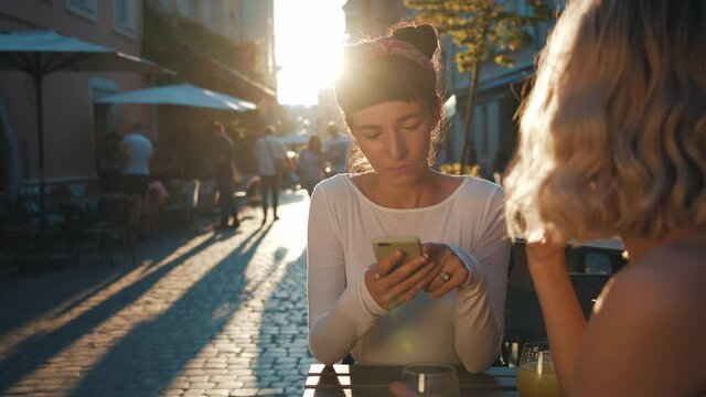 Two serene young woman spending sunny evening while sitting at the street outdoor cafe, drinking juice, showing photos, scrolling website or article in the internet communicate and sharing news