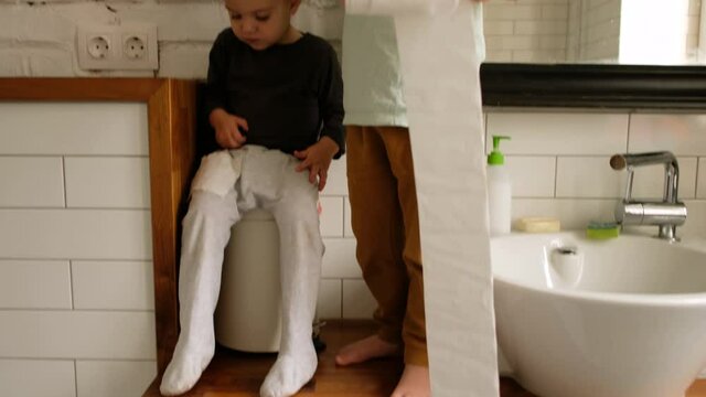 Cute little boy and toddler sister play with toilet paper standing on wooden countertop at vessel sink and mirror in bathroom