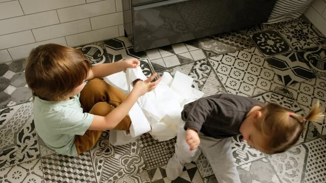 Cute blond little kid with playful sister has fun with uncoiled white toilet paper on tiled floor in contemporary bathroom