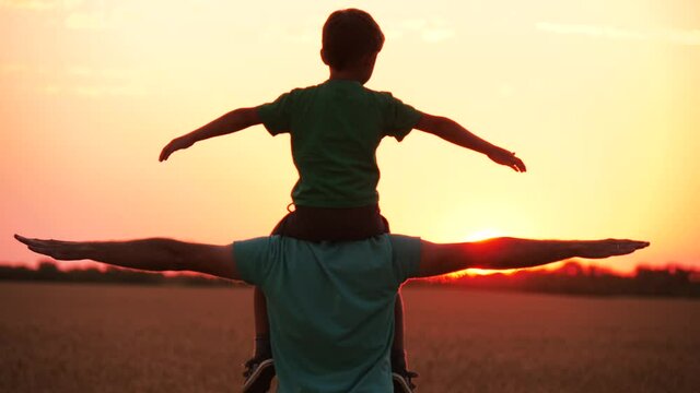 A father holds his son on his shoulders, arms outstretched, playing airplane flight. Teamwork, happy family at sunset.