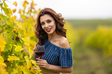 Naklejka na ściany i meble A beautiful woman with a glass of red wine in the vineyard on autumn day.