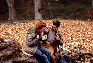 Two young hikers with backpack sitting on collapsed trunk resting and drinking tea after walking in forest.