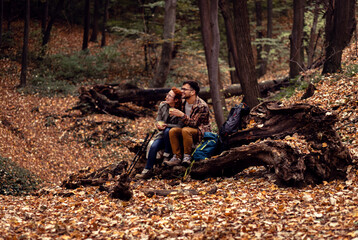 Two young hikers with backpack sitting on collapsed trunk resting and drinking tea after walking in forest.