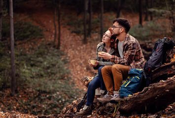 Two young hikers with backpack sitting on collapsed trunk resting and drinking tea after walking in forest.