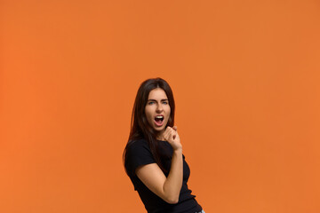 I am a popular singerJoyful caucasian woman in black t-shirt holds hand near mouth as microphone, sings songs in karaoke, being in happy mood. Isolated over an orange background.