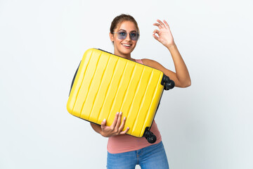 Obraz premium Young student woman isolated on white background in vacation with travel suitcase and making OK sign