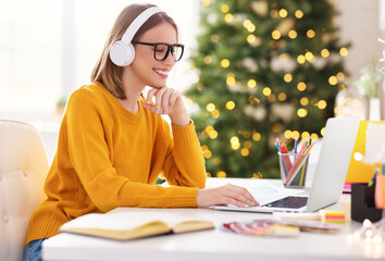 Happy young woman freelancer in headphones working remote at laptop at home before Christmas.