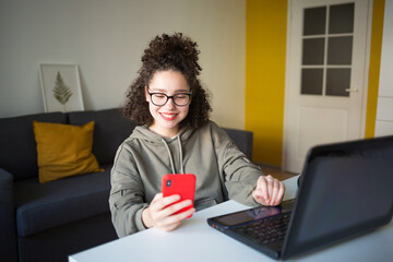 happy mixed rase girl sits at computer screen laptop and looks at phone. student talks over...