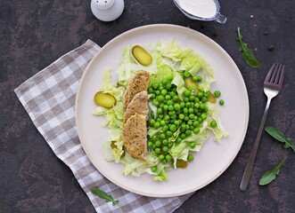 Healthy salad made of iceberg lettuce, fresh green peas, pickled cucumbers and pork on a light ceramic plate on a dark brown concrete background. Salad recipes. Healthly food.