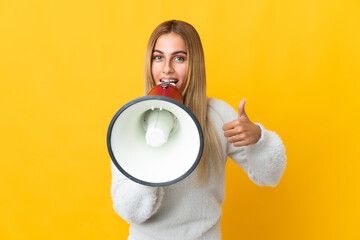 Young blonde woman isolated on yellow background shouting through a megaphone to announce something and with thumb up