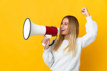 Young blonde woman isolated on yellow background shouting through a megaphone to announce something...