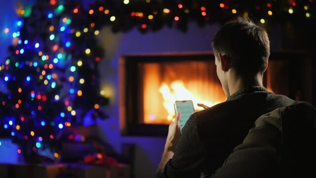 Young man uses a smartphone by the fireplace decorated for Christmas. Gift order