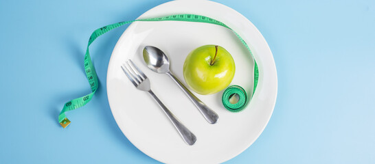 Top view spoon and fork, green apple on white ceramic plate with green Measuring tape on blue  background. dieting, weight loss, obesity and food control concept